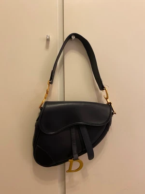 Marinblå Dior Saddle Bag i läder - Marinblå Dior Saddle Bag i slätt läder med guldfärgade detaljer. Väskan kommer med både kort och längre axelrem samt magnetstängning. Mycket bra skick.