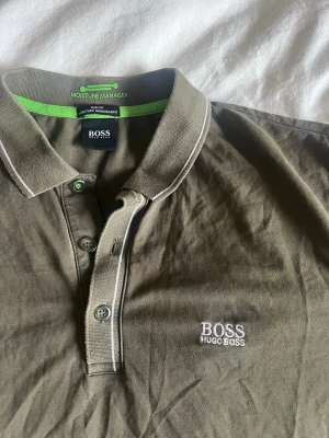 Olivgrön pikétröja från Hugo Boss - Snygg olivgrön pikétröja från Hugo Boss med vit logga broderad på bröstet. Tröjan har klassisk krage, knappar framtill och diskreta vita detaljer längs kragen och knappslån. Tillverkad i bomull och har en relaxed fit som passar perfekt till vardagslooken.