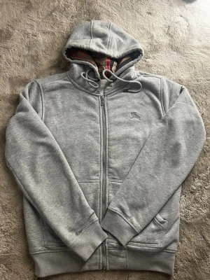 Grå zip-hoodie från Burberry - Snygg grå hoodie med dragkedja från Burberry. Klassisk huva med dragsko och ikoniskt rutigt foder. Broderad logga på bröstet och två fickor framtill. Perfekt för en chill och stilren look.