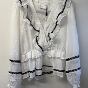 Vit volangblus med svarta detaljer - Söt vit blus från NEO NOIR med volanger och svarta spetsdetaljer längs v-ringningen, ärmarna och nederkanten. Blusen har lång ärm med resår vid ärmslut och är tillverkad i ett lätt, luftigt tyg som ger en romantisk vibe.