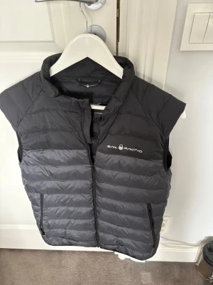 Mörkgrå dunväst från Sail Racing - Snygg mörkgrå dunväst från Sail Racing med quiltad design och dragkedja framtill. Västen har två praktiska fickor med dragkedja och en hög krage. Perfekt för lager-på-lager och stilren look.