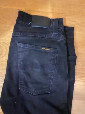 Svarta jeans från Nudie Jeans - Säljer ett par svarta jeans från Nudie Jeans med klassisk femficksdesign och smal passform. Jeansen har knappgylf och subtila sömdetaljer på bakfickan. Tillverkade i slitstark denim, perfekta för en stilren vardagslook. Storlek 30 men sitter som 31