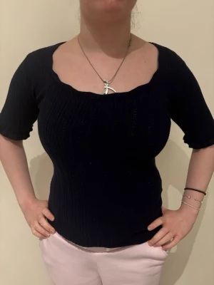 Mörkblå ribbad topp med kort ärm - Snygg mörkblå ribbad topp med korta ärmar och unik, vågig urringning. Figurnära passform som framhäver siluetten. Perfekt till jeans eller kjol för en trendig look. Materialet känns mjukt och stretchigt.