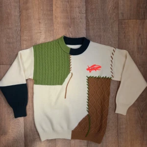 Patchwork stickad tröja med mönster - Unik stickad tröja i patchwork-stil med färgblock i grön, brun, marinblå och offwhite. Tröjan har olika stickade strukturer, kontrastsömmar och en röd broderad detalj framtill. Rund halsringning och långa ärmar. Perfekt för dig som gillar statement-plagg.