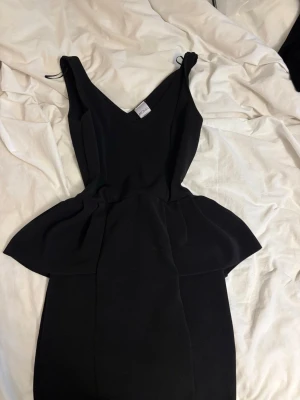 Svart peplumklänning från Monki - Snygg svart klänning från Monki i peplum-modell med v-ringning och ärmlös design. Perfekt för dig som vill ha en stilren och trendig look. Klänningen har en figurnära passform och markerad midja som ger en snygg siluett.
