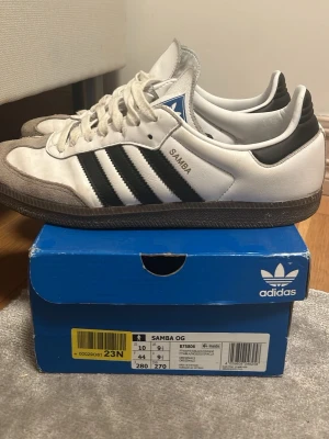 Adidas Samba skor storlek 44 - Klassiska Adidas Samba sneakers i vitt läder med svarta detaljer och de ikoniska tre ränderna på sidan. Grå mocka vid tån och brun gummisula. Blå Adidas-logga på plösen och SAMBA-tryck i guld på sidan. Perfekta för en sportig och retro look.