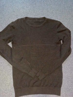 Mörkbrun stickad tröja - En enkel och stilren mörkbrun stickad tröja med rund halsringning och långa ärmar. Tröjan har ribbade muddar vid ärmslut och nederkant. Perfekt att matcha med jeans eller kjol för en avslappnad look.