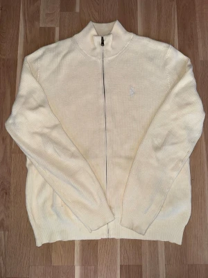 Krämvit ribbstickad zip-kofta från Polo Ralph Lauren - Elegant och tidlös zip-kofta i krämvit/off-white nyans från Polo Ralph Lauren. Ribbstickad struktur med broderad logga på bröstet och hel dragkedja. Perfekt för en clean, preppy look – lika snygg till jeans som över skjorta.  ✔️ Klassisk och exklusiv modell ✔️ Hel dragkedja ✔️ Diskret broderad logga ✔️ Perfekt lager-på-lager ✔️ Funkar året runt  Mycket stilren färg som lyfter hela outfiten.  En riktig garderobsklassiker – först till kvarn.
