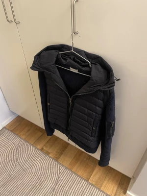 Mörkblå cardigan från Moncler - Snygg mörkblå hybridjacka från Moncler med quiltad front och stickade ärmar. Jackan har huva, dragkedja framtill och Moncler-logga på ärmen. Perfekt för dig som vill ha både stil och funktion i ett plagg.