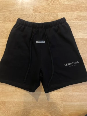 Svarta shorts Essentials Fear of God - Säljer ett par svarta shorts från Essentials Fear of God med tryckt logga på benet och snörning i midjan. De har en relaxed fit och är perfekta för sommaren. Tillverkade i mjuk bomull för maximal komfort.