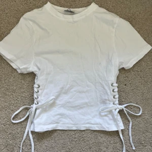 Vit t-shirt med snörning i sidorna - Säljer en vit kortärmad topp med cool snörningsdetalj i båda sidorna. Använd ca 3ggr säljer pga att den inte används, köpte på asos🥰