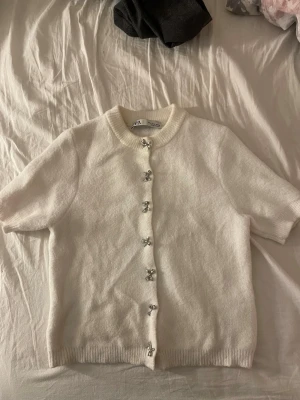 Vit stickad kortärmad  tröja ZARA - Vit stickad tröja från ZARA med korta ärmar och ribbad krage. Koftan har dekorativa silvriga knappar framtill och är gjord i ett mjukt material. Passar även på mig som är en xs