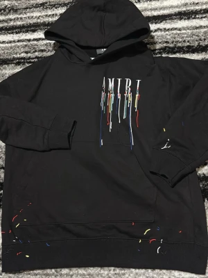 Svart hoodie från Amiri  - Säljer en svart hoodie från Amiri med huva och känguruficka. Framsidan har AMIRI-logga med broderade färgstänk i rött, blått, gult och vitt som rinner ner. Det finns även små färgstänk på ärm och nederkant. Perfekt för dig som gillar streetwear och unik design.