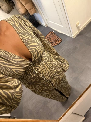 Zebramönstrad långklänning från Adoore - Säljer en zebramönstrad långklänning från Adoore med v-ringning, markerad midja och dekorativa knappar framtill. Klänningen har långa puffärmar och dragkedja i sidan. Färgerna går i beige och mörkbrunt, vilket ger en trendig look.