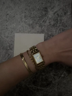 Guldfärgad rektangulär klocka - Stilren klocka med rektangulär vit urtavla och guldfärgad länk i metall. Klassisk design med tydliga svarta siffror och visare. Perfekt accessoar för dig som gillar tidlös och elegant stil.