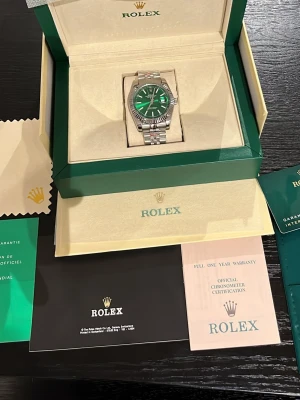 Rolex klocka med grön urtavla - Exklusiv Rolex klocka med grön urtavla och silverfärgat armband i rostfritt stål. Klockan har klassisk rund form, räfflad bezel och tydliga indexmarkeringar. Levereras i originalbox med certifikat och tillbehör.