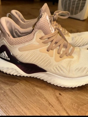 Beige Adidas Alphabounce sneakers - Snygga Adidas Alphabounce sneakers i beige och ljusrosa med vita sulor och mörklila detaljer. Ovandelen är i mesh för bra andningsförmåga och skorna har klassiska snören. Perfekta för dig som vill ha både stil och komfort.