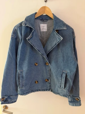 Blå dubbelknäppt jeansjacka Lindex - Snygg blå jeansjacka från Lindex med dubbelknäppning och stora bruna knappar. Jackan har två framfickor, bred krage och justerbara ärmslut. Klassisk modell i jeansmaterial som ger en avslappnad och trendig look.