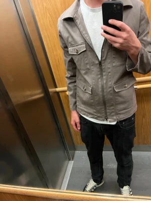 Beige overshirt från John Henric - Snygg beige overshirt från John Henric med dragkedja framtill och fyra fickor med lock. Jackan har en rak passform och klassisk krage, perfekt att slänga över en t-shirt.