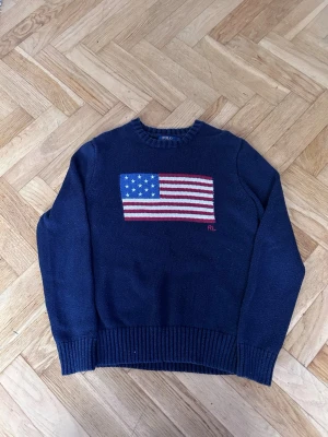 Mörkblå stickad tröja Polo Ralph Lauren - Mörkblå stickad tröja från Polo Ralph Lauren med amerikansk flagga i rött, vitt och blått på bröstet. Rund halsringning och ribbstickade muddar vid ärmslut och nederkant. RL-broderi i rött vid flaggan.