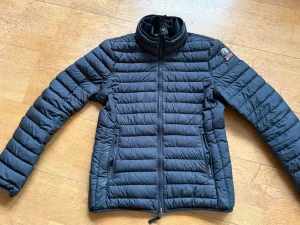 Svart dunjacka från Parajumpers - Snygg svart dunjacka från Parajumpers med quiltad design och hög krage. Jackan har dragkedja framtill, två sidofickor med dragkedja och en patch på ena ärmen. Perfekt för kalla vinterdagar och riktigt lätt att bära.