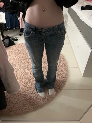 Skit snygga Miss Me jeans - Säljer ett par blå Miss Me jeans med låg midja och bootcut. Jeansen har snygga slitningar och detaljer som broderad kors på ena bakfickan samt nitar och rivet tyg på andra bakfickan. Det är svart tyg under hålet. Materialet är klassisk jeans med stretch och cool faded effekt. Innerbenslängd 79 cm, midjemått 38 cm.