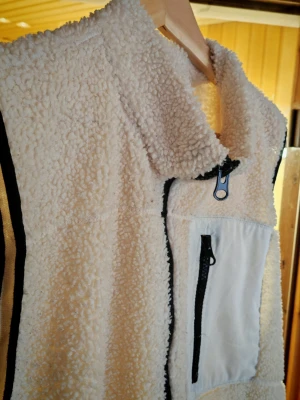 Vit teddyjacka med dragkedja - Skön vit teddyjacka i fluffigt material med svart dragkedja och vit bröstficka med svart blixtlås. Perfekt för vinter eller vår. Lager 157