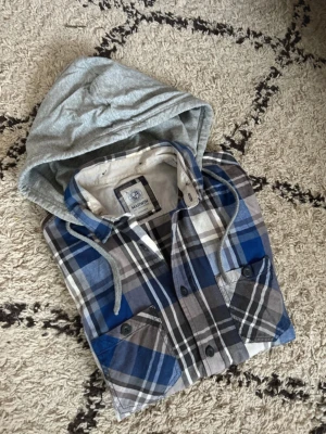 Rutig skjorta med grå luva från Marwin Archive - Säljer en rutig skjorta från Marwin i blå, grå, svart och vit med en grå luva och snörning. Skjortan har knappar framtill och två bröstfickor. Perfekt för en avslappnad Archive streetstyle-look.