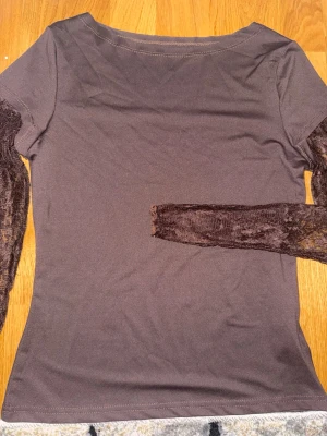 Brun topp med spetsärmar - Snygg brun topp med transparenta spetsärmar som ger en unik touch. Modellen är figurnära och har rund halsringning. Perfekt för dig som vill ha något stilrent men ändå med en twist.