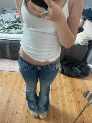 Blå bootcut jeans med låg midja - Säljer ett par blå bootcut jeans med låg midja och slitningar framtill. Jeansen har ljusare tvättade partier på låren. Perfekta för en avslappnad och trendig look.