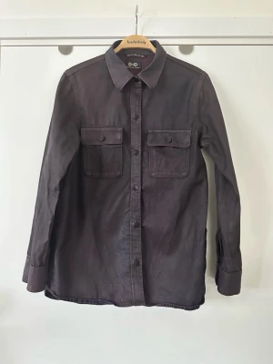 Mörklila skjorta från D-ID - Stilren mörklila skjorta från D-ID med två bröstfickor med lock och knappar. Skjortan har klassisk krage, långa ärmar och knäppning framtill. Perfekt för dig som gillar en avslappnad men snygg look. Storlek S för herr, storlek M för dam. 