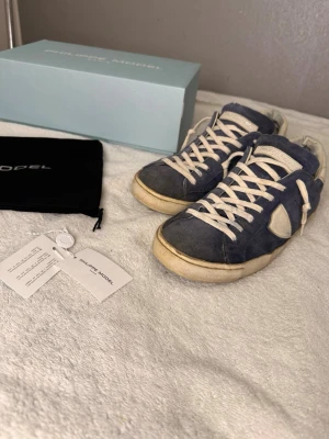 Blå Philippe Model sneakers i mocka - Snygga blå sneakers från Philippe Model i mocka med vita detaljer och vita skosnören. Klassisk låg modell med vit sula och ikonisk sköld på sidan. Perfekta för en trendig och avslappnad stil.