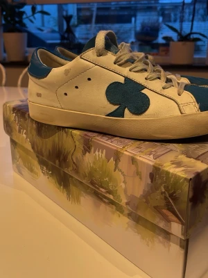 Vit och blå sneaker från The Gallerist - Sneaker i vitt läder med blå detaljer och en stor blå klöver på sidan. Skon har vita skosnören, blå häl och plös samt svart sula undertill. Snygg och unik design som sticker ut.