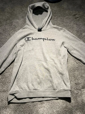Grå Champion hoodie med logga - Snygg grå hoodie från Champion med stor svart logga över bröstet. Klassisk modell med huva och känguruficka framtill. Mjuk bomullsblandning som är skön att ha på sig. Perfekt för en avslappnad streetstyle-look.