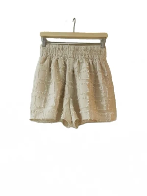 Beige shorts med fransig struktur - Unika beige shorts med hög midja och elastisk midjeresår. Shortsen har ett rutigt, fransigt mönster som ger en cool och trendig look. Perfekta för varma dagar och lätta att matcha med olika toppar.