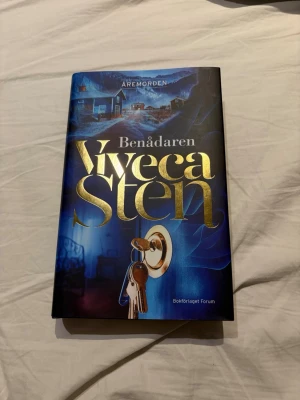 Benåadaren av Viveca Sten - En spännande roman i serien Åremorden av Viveca Sten. Omslaget är blått med guldtext och motiv av nycklar och ett hus. Boken är inbunden och har en mystisk och modern design som lockar till läsning. Den är helt ny och aldrig läst. Stor inbunden bok, inte en klassisk coffe table-bok.