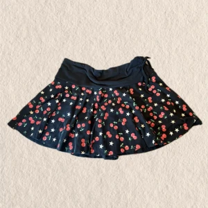 Svart kortkjol med körsbär från Akama - Snygg svart kortkjol från Akama med körsbärs- och stjärnmönster. Kjolen har bred midjeresår och knytband i midjan för extra passform. Perfekt för dig som gillar lekfulla detaljer och vill sticka ut.