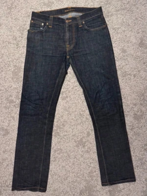 Mörkblå  jeans från Nudie  W30/L30 -  Säljer ett par riktigt snygga dry denim-jeans från NUDIE  i mörk rå denim med dåm klassiska orangea sömmarna . Passformen är clean och sitter sjukt bra på kroppen, perfekt för dig som kör grich-stil och vill ha ett par jeans som lyfter hela outfiten. Den här typen av denim formar sig efter dig med tiden och kommer ge galet feta fades ju mer du använder dem. Ett måste i garderoben för dig som gillar stilrena men ändå råa plagg. Jeansen är i fint skick med bara naturligt