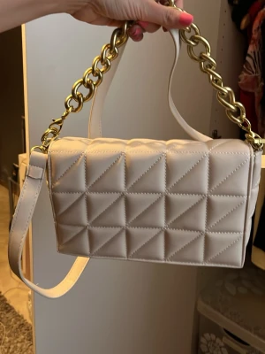 Beige quiltad handväska med guldkedja - Säljer en beige quiltad handväska med rektangulär form och stora guldfärgade kedjedetaljer. Väskan har både axelrem och handtag, och är gjord i ett mjukt, vadderat material som ger en lyxig känsla. Perfekt accessoar för att lyfta din outfit.