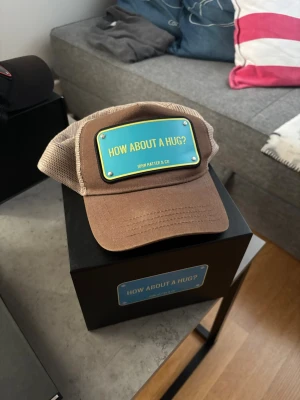 Brun truckerkeps John Hatter & Co - Säljer en brun truckerkeps från John Hatter & Co med mesh på sidorna och bak. Framtill sitter en blå skylt med texten 'HOW ABOUT A HUG?'. Kepsen har böjd skärm och justerbar snapback. Perfekt för dig som vill sticka ut med en cool accessoar.