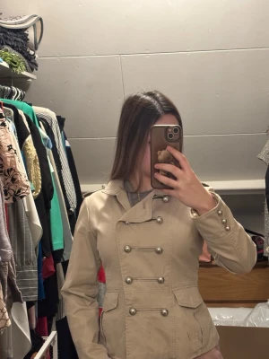Beige trenchcoat med dubbelknäppning  - Snygg beige trenchcoat med klassisk dubbelknäppning och dekorativa silverfärgade knappar. Jackan har axelklaffar, fickor med lock och ett slitsat parti bak med knappar. Perfekt för dig som vill ha en stilren och tidlös look. Från Famale collection storlek S