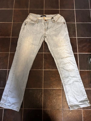 Ljusblå raka/ bootcut jeans - Säljer ett par ljusblå jeans med raka/ bootcut ben och klassisk femficksmodell. Jeansen har ljusa slitningar framtill och detaljerade bakfickor. Tillverkade i denim med kontrastsömmar och knappgylf.