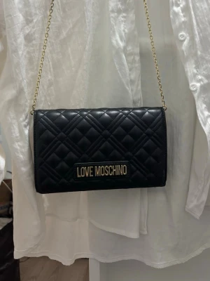 Svart quiltad axelväska Love Moschino - Snygg svart axelväska från Love Moschino i quiltat konstläder med guldfärgad kedja och logotyp framtill. Väskan har en stilren, rektangulär form och passar perfekt för att lyfta din outfit med en trendig touch, funkar till vardags eller för en speciell dag.