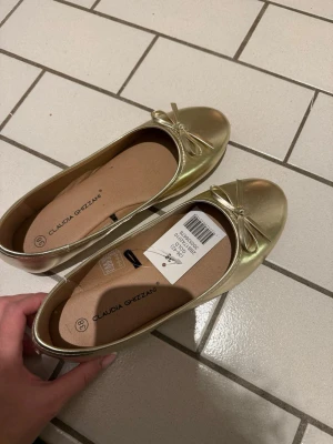 Guldiga ballerinaskor Claudia Ghizzani - Snygga ballerinaskor i metallic guld från Claudia Ghizzani. Skorna har rund tå, liten rosett framtill och en klassisk, enkel design. Tillverkade i ett glansigt syntetmaterial med beige innersula för extra komfort.