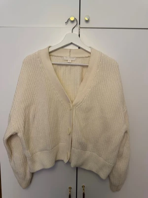 Krämvit stickad kofta från H&M - Mysig krämvit stickad kofta från H&M med v-ringning och stora knappar framtill. Koftan har en avslappnad passform, ribbade muddar och är gjord i ett tjockare garn som känns riktigt skönt. Perfekt att slänga över en t-shirt eller topp.