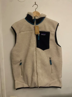 Beige fleeceväst från Patagonia - Mysig beige fleeceväst från Patagonia med blå detaljer, hög krage och dragkedja framtill. Västen har två fickor med dragkedja och en bröstficka i mörkblått tyg. Perfekt för lager-på-lager och outdoor vibes.