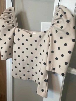 Beige topp med puffärm och prickar - Trendig beige topp med svarta prickar och puffiga, korta ärmar. Fyrkantig urringning och figurnära passform ger en cool retrovibe. Perfekt att styla med jeans eller kjol för en snygg look.