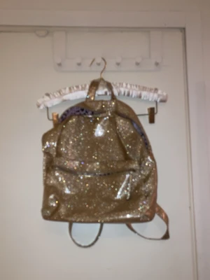 Glittrig guldryggsäck - Säljer en riktigt snygg ryggsäck i glittrigt guldigt material. Väskan har justerbara axelband, ett stort huvudfack och en mindre ficka framtill. Perfekt för dig som vill sticka ut och älskar glitter! Materialet känns plastigt och väskan har en cool, ungdomlig vibe.