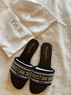 Svarta sandaler från Christian Dior - Snygga svarta sandaler från Christian Dior med band och vit broderad logotyp. Sandalerna har en klassisk slip-on design och är tillverkade i textil och läder. Perfekta för att ge din outfit en lyxig touch.