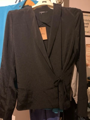 Svart dam kavaj från J.Lindrsberg med knytband. - Stilren svart kavaj från J Lindesberg med knytband i midjan och klassisk krage. Kavajen har lång ärm och en lätt glansig . Liten i storleken Perfekt för dig som vill ha en enkel men snygg look.